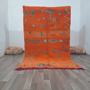 May include: A hand-woven Moroccan rug with a bright orange background and intricate geometric patterns in shades of blue, green, and yellow. The rug has a fringe border and is made of wool.