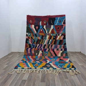 May include: A colorful, geometric patterned rug with a textured surface. The rug features a variety of colors including red, blue, green, and yellow. The rug has a fringe along the bottom edge and is displayed on a wooden floor.