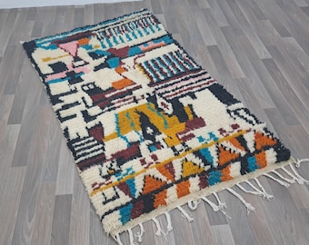Op de afbeelding: Een kleurrijk, geometrisch patroon tapijt met een witte achtergrond en een franje rand. Het tapijt heeft tinten van blauw, oranje, geel, rood en zwart.