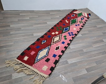 May include: A long, narrow rug with a geometric pattern in shades of red, pink, white, black, blue, and green. The rug has fringe on both ends.