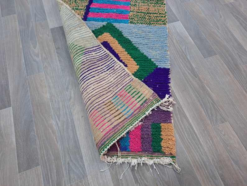 May include: A colorful, geometric patterned rug with a fringed edge. The rug is folded in half, revealing the back side, which is a beige color with a striped pattern.