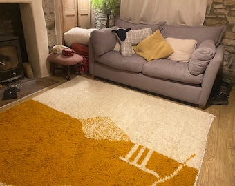 May include: A white and mustard yellow shag rug with a geometric design. The rug is in a living room with a gray sofa and a wooden coffee table.