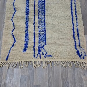 May include: A white and blue Moroccan rug with a fringe border. The rug has a geometric pattern with blue lines and zigzags on a white background.