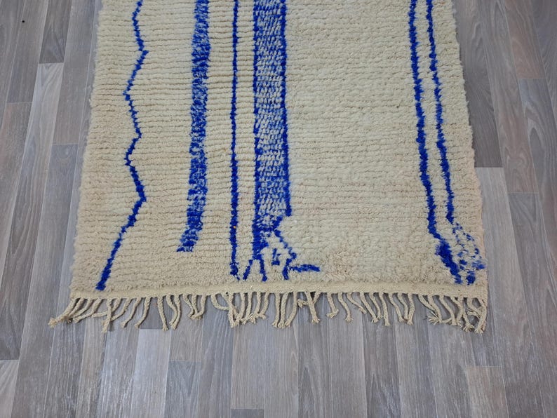 May include: A white and blue Moroccan rug with a fringe border. The rug has a geometric pattern with blue lines and zigzags on a white background.