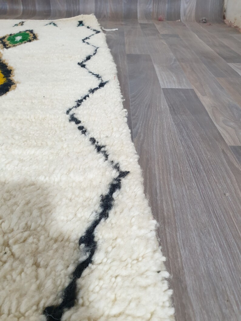May include: A close-up of a white, textured rug with a black zigzag pattern and colorful geometric shapes. The rug has a thick pile and a soft appearance, suitable for home decor.