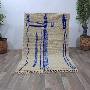 May include: A white wool rug with blue geometric designs. The rug has a fringe border and is laid out on a wooden floor.