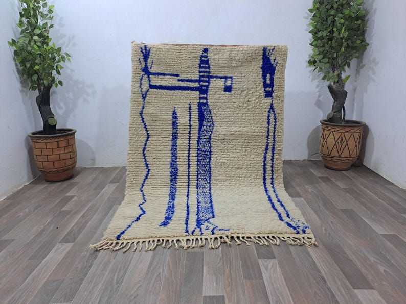 May include: A white wool rug with blue geometric designs. The rug has a fringe border and is laid out on a wooden floor.