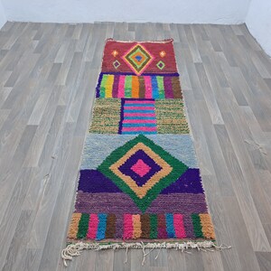 May include: A colorful Moroccan rug with geometric patterns in shades of red, green, blue, pink, and brown. The rug has a fringed edge and is made of wool.