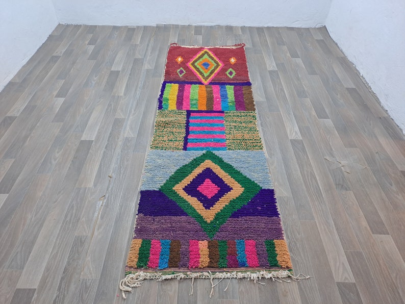 May include: A colorful Moroccan rug with geometric patterns in shades of red, green, blue, pink, and brown. The rug has a fringed edge and is made of wool.