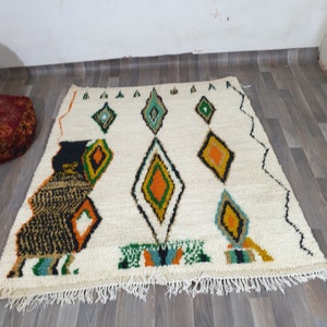 May include: Cream-colored, rectangular rug with a geometric diamond pattern in green, orange, and black. The rug has a fringed edge and a stylized animal figure on the left side. The rug is placed on a wooden floor.