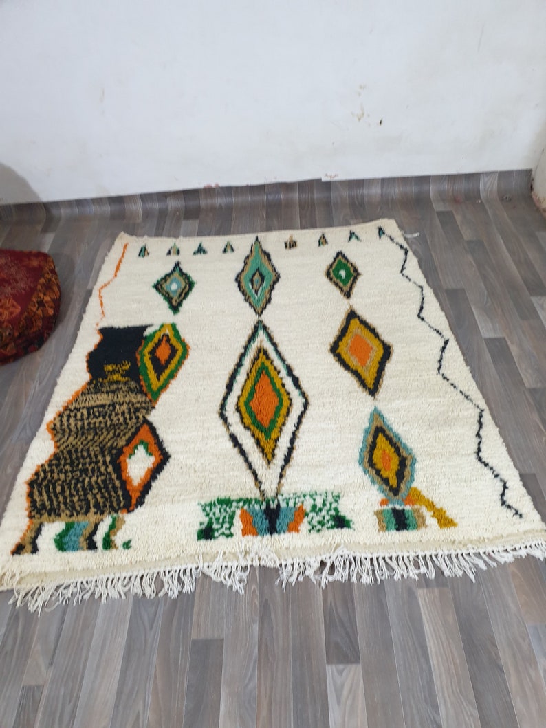 May include: Cream-colored, rectangular rug with a geometric diamond pattern in green, orange, and black. The rug has a fringed edge and a stylized animal figure on the left side. The rug is placed on a wooden floor.