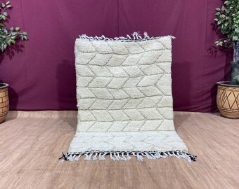May include: A white and cream Moroccan rug with a geometric pattern and fringe. The rug is laid out on a wooden floor.