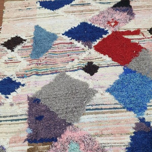 May include: A colorful, handwoven rug with a patchwork design. The rug features various geometric shapes in shades of blue, red, pink, gray, and brown. The rug is made with a variety of textures, including fluffy and woven.