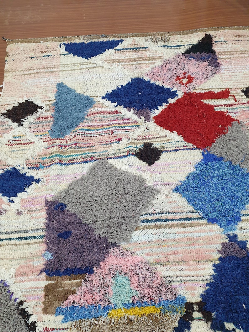 May include: A colorful, handwoven rug with a patchwork design. The rug features various geometric shapes in shades of blue, red, pink, gray, and brown. The rug is made with a variety of textures, including fluffy and woven.