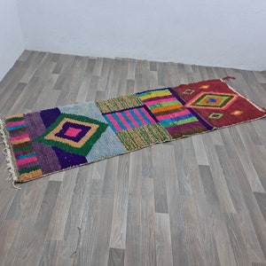 May include: A colorful, geometric patterned rug with a mix of pink, green, blue, purple, and yellow. The rug is made of woven fabric and has a fringed edge.