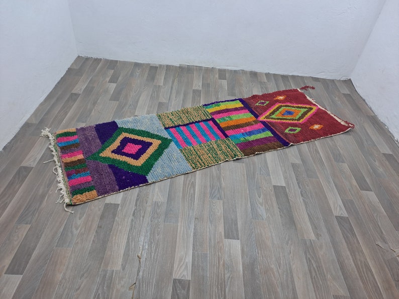 May include: A colorful, geometric patterned rug with a mix of pink, green, blue, purple, and yellow. The rug is made of woven fabric and has a fringed edge.