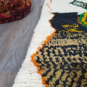 May include: A portion of a white, black, and orange patterned rug with a shaggy texture. A round, red and orange patterned ottoman is in the background. The rug has a geometric design with a mix of colors and textures.