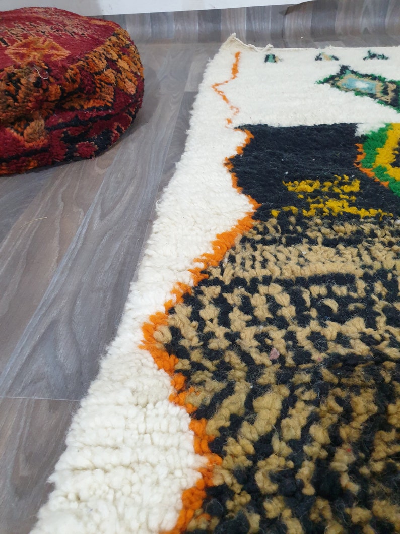May include: A portion of a white, black, and orange patterned rug with a shaggy texture. A round, red and orange patterned ottoman is in the background. The rug has a geometric design with a mix of colors and textures.