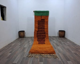 May include: A long, rectangular rug with a geometric design. The rug is orange with a green border and a brown building design in the center.