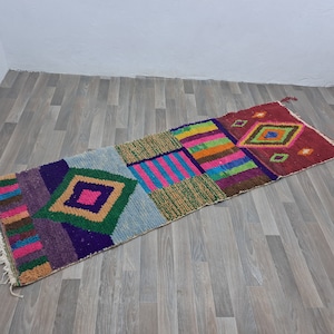 May include: A colorful, geometric patterned rug with a mix of pink, blue, green, purple, and brown. The rug is made of woven fabric and has a fringed edge.
