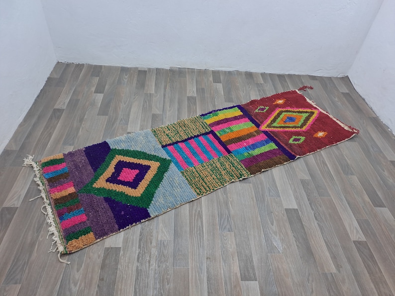 May include: A colorful, geometric patterned rug with a mix of pink, blue, green, purple, and brown. The rug is made of woven fabric and has a fringed edge.