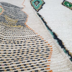 May include: A folded white rug with geometric patterns. The rug features black, orange, and yellow stripes and dots on one side, and diamond shapes in teal and green on the other. The rug has a textured surface.