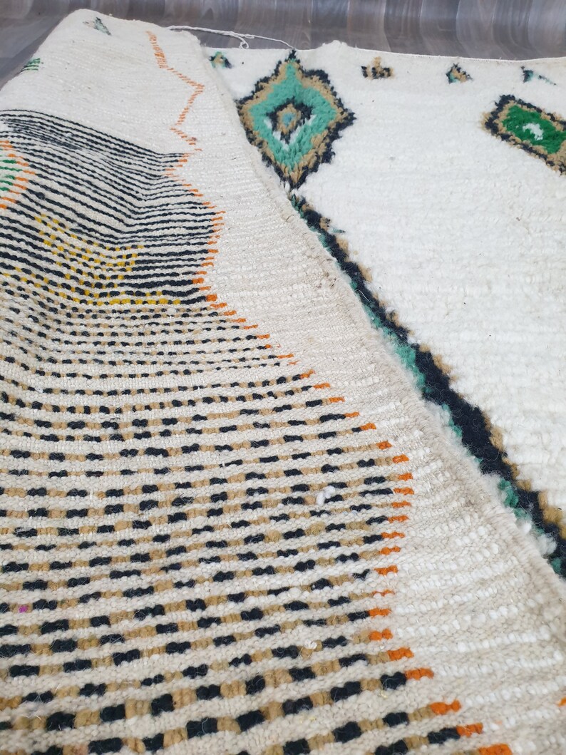 May include: A folded white rug with geometric patterns. The rug features black, orange, and yellow stripes and dots on one side, and diamond shapes in teal and green on the other. The rug has a textured surface.
