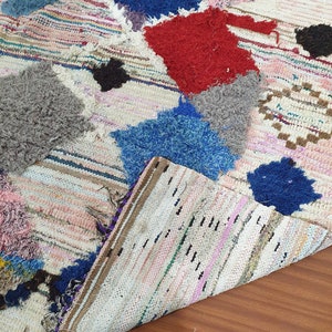 May include: A white, handwoven rug with colorful patches of fabric sewn on top. The patches are in shades of blue, red, pink, and gray. The rug is folded in half, showing the underside.
