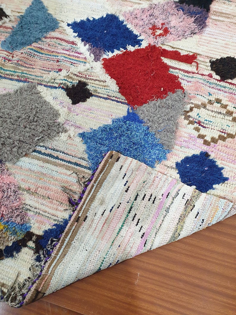 May include: A white, handwoven rug with colorful patches of fabric sewn on top. The patches are in shades of blue, red, pink, and gray. The rug is folded in half, showing the underside.