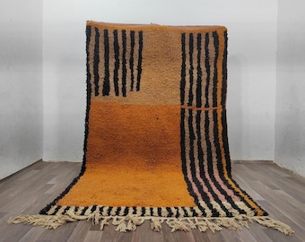 May include: A rectangular area rug featuring a geometric colour-blocked design in shades of orange, tan, and black. The rug displays vertical black stripes on the sides and a solid orange section at the bottom, finished with cream-coloured fringe.