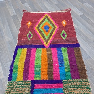 May include: A colorful Moroccan rug with a geometric pattern in shades of red, orange, yellow, green, blue, and purple. The rug has a diamond shape in the center and is made of wool.