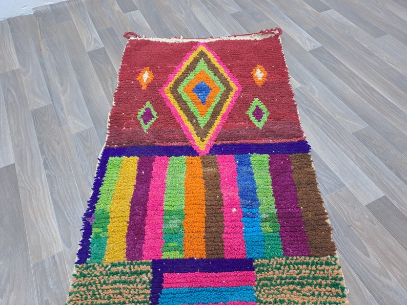 May include: A colorful Moroccan rug with a geometric pattern in shades of red, orange, yellow, green, blue, and purple. The rug has a diamond shape in the center and is made of wool.