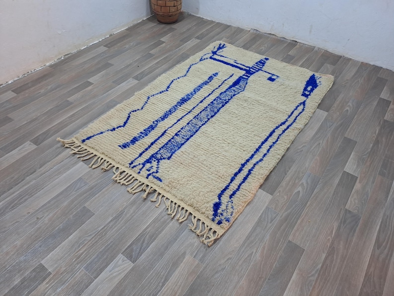 May include: A white and blue Moroccan rug with a geometric design. The rug has a fringe border and is made of wool.