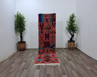 May include: A long, rectangular runner rug with a dusty pink base colour, featuring a variety of geometric and abstract symbols in vibrant shades of blue, orange, green, and dark purple. The rug has a knotted texture and white fringe at the bottom.