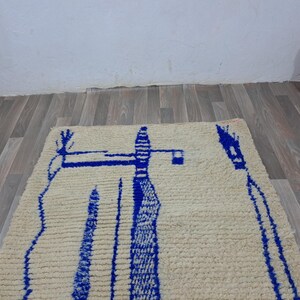 May include: A white wool rug with a blue abstract design. The design features a tall, thin shape with a smaller shape on top. The rug has a fringe border.