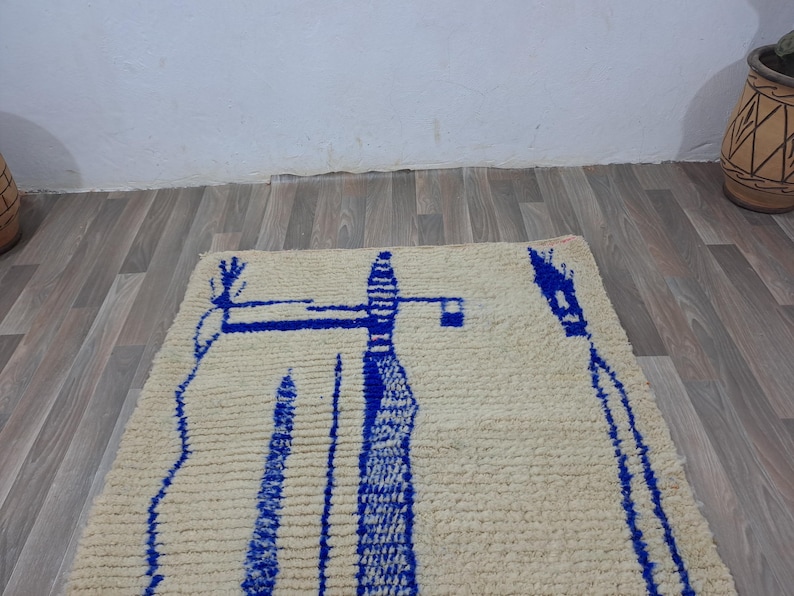 May include: A white wool rug with a blue abstract design. The design features a tall, thin shape with a smaller shape on top. The rug has a fringe border.
