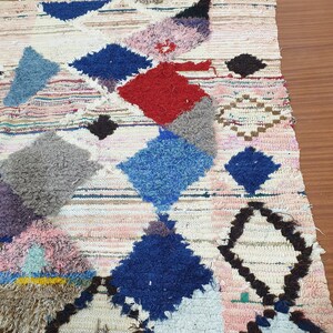 May include: A white, handwoven rug with a geometric pattern in shades of blue, red, pink, and brown. The rug has a fringe border.