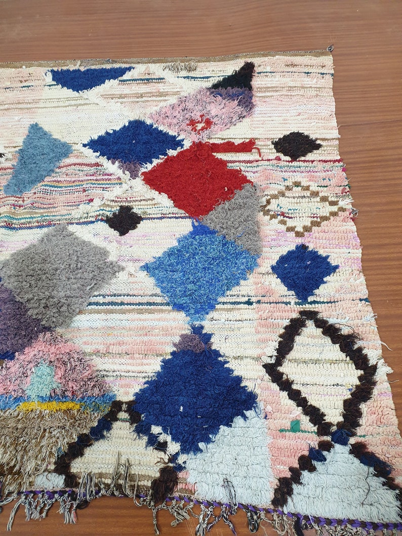 May include: A white, handwoven rug with a geometric pattern in shades of blue, red, pink, and brown. The rug has a fringe border.