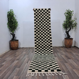 Op de afbeelding: Een zwart-wit geblokt tapijt met groene accenten. Het tapijt heeft een franje rand en is gemaakt van wol.