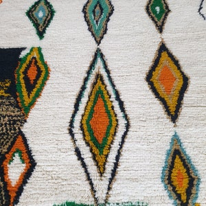 May include: A cream-colored, textured rug with a repeating diamond pattern. The diamonds are outlined in black and filled with various colors, including orange, green, yellow, and teal. The rug has a thick, plush pile.