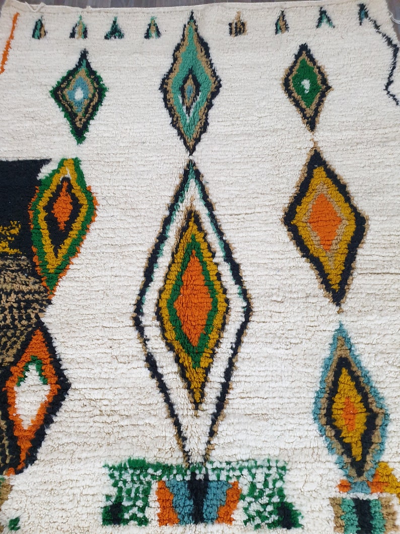 May include: A cream-colored, textured rug with a repeating diamond pattern. The diamonds are outlined in black and filled with various colors, including orange, green, yellow, and teal. The rug has a thick, plush pile.
