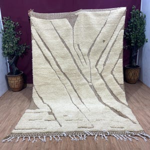 Peut inclure: Un tapis blanc et crème avec un motif géométrique. Le tapis a une bordure en franges et est fait de laine.