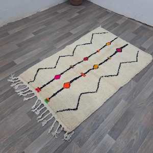 Peut inclure: Un tapis rectangulaire blanc cassé orné d'un motif de lignes noires en zigzag et de lignes droites, agrémenté de petits motifs en forme de losange aux couleurs orange, rose, jaune et vert. Le tapis est terminé par des franges blanches.