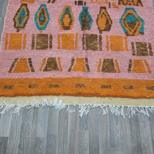 May include: A pink, orange, and brown patterned rug with a fringe border. The rug has a geometric design with a repeating pattern of squares and triangles.