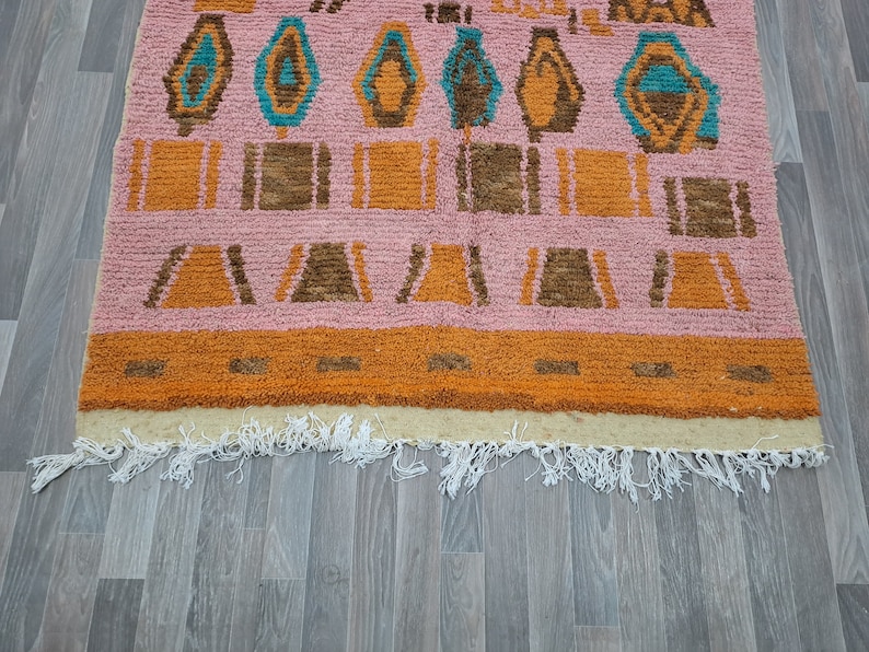 May include: A pink, orange, and brown patterned rug with a fringe border. The rug has a geometric design with a repeating pattern of squares and triangles.