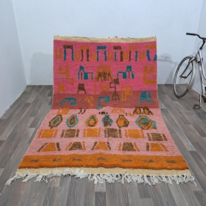 May include: A pink, orange, and blue Moroccan rug with geometric patterns. The rug has a fringe border and is made of wool.