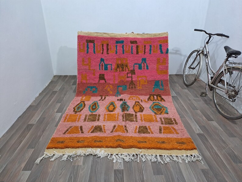 May include: A pink, orange, and blue Moroccan rug with geometric patterns. The rug has a fringe border and is made of wool.