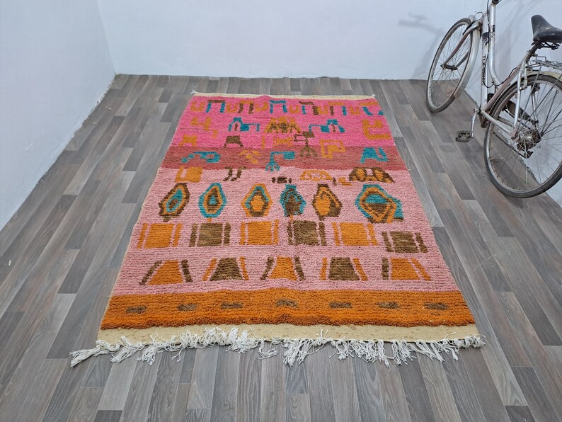 May include: A colorful Moroccan rug with a geometric pattern in shades of pink, orange, brown, and blue. The rug has a fringe border and is made of wool.