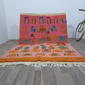 May include: A pink and orange Moroccan rug with a geometric pattern. The rug has a fringe border and is made of wool.