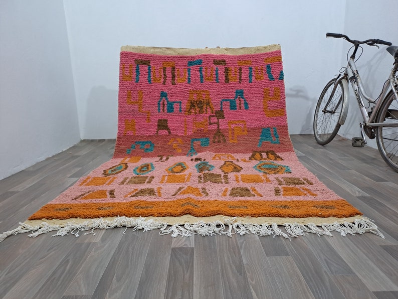 May include: A pink and orange Moroccan rug with a geometric pattern. The rug has a fringe border and is made of wool.